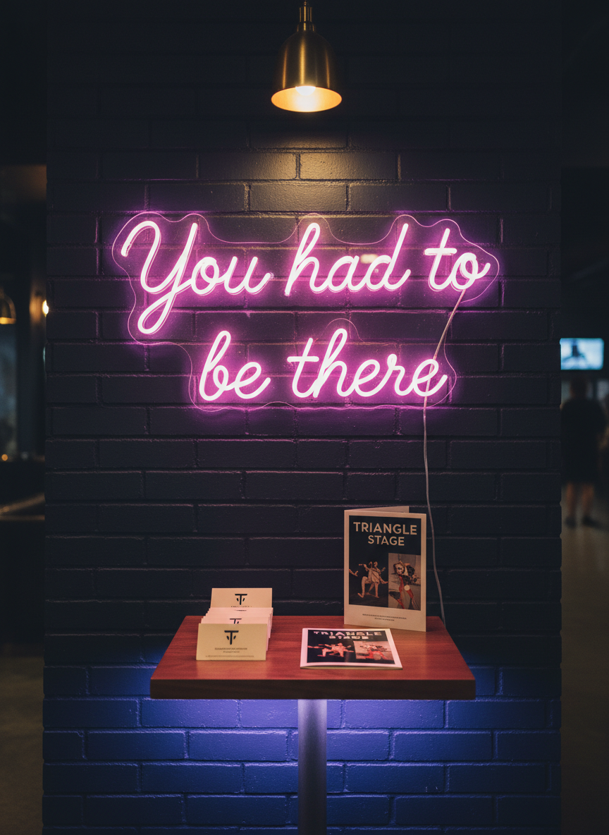 A handwritten “You had to be there” neon-style sign rendered in glowing, curved tubing of electric pink and soft violet, mounted against an exposed brick wall painted deep midnight blue. Below it, a small table holds a neat stack of minimalist membership info cards and a glossy, folded season brochure for a theater company. Subtle uplighting from hidden LED strips under the table adds a modern, playful glow, while a single overhead pendant casts a gentle pool of warm light on the printed materials. Captured straight-on with symmetrical framing and photographic realism, the background falls slightly out of focus. The atmosphere feels conspiratorial and lively, evoking a secret invitation to late-night arts experiences in the Triangle.